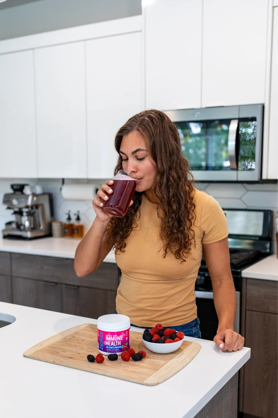 Woman drinking a glass of Zipfizz Immune Health in a kitchen with a Zipfizz Immune Health pack and berries on the counter.